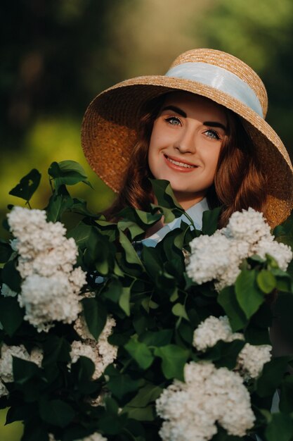 Een stijlvolle vrouw in een strooien hoed poseert op lila bloemen in een zonnig voorjaarspark. rustig portret van een mooi meisje dat zich met een boeket van seringen in de lentetuin bevindt.