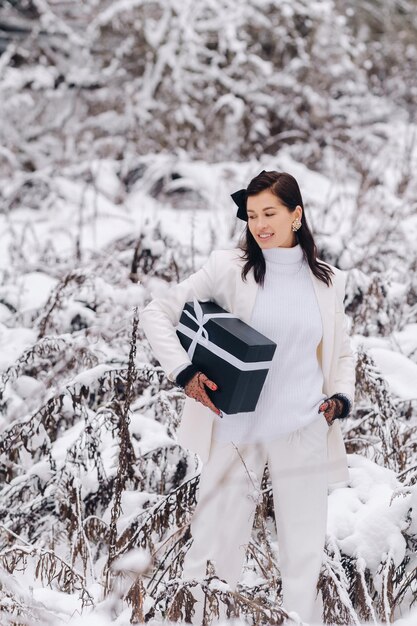 Een stijlvolle vrouw met een wit pak met een nieuwjaarscadeau in haar handen in een winterbos Een meisje in de natuur in een besneeuwd bos met een geschenkdoos