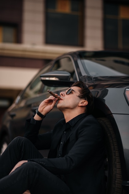 Een stijlvolle zakenman rookt sigaren in de buurt van een luxeauto. Mode en zaken.
