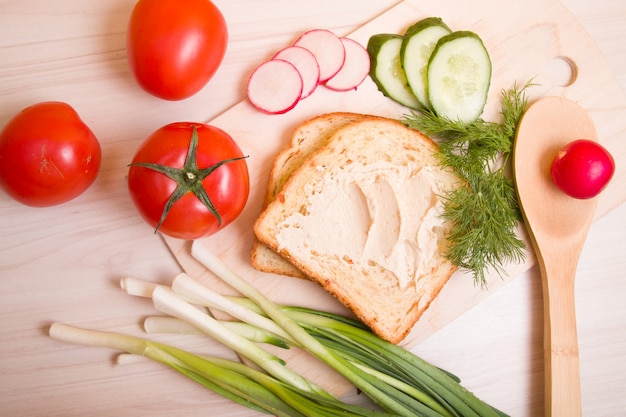 Een stuk brood uitgespreid met hummus, groenten en dille op een houten tafel