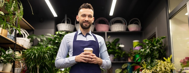 Een succesvolle bloemist met een kopje koffie in zijn handen staat in een bloemenwinkel en kijkt naar de