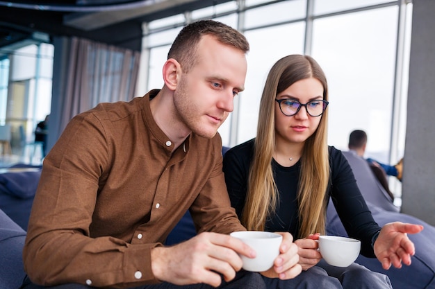 Een succesvolle jonge vrouw manager met haar baas zit met een laptop op een gestoffeerde fauteuil bij het panoramische raam en drinkt koffie. Zakenvrouw en man werken aan een nieuw project