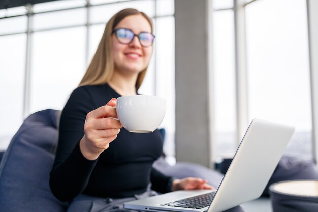 Een succesvolle jonge vrouwelijke manager zit met een laptop op een zachte fauteuil bij het panoramische raam en drinkt koffie. Zakenvrouw die aan een nieuw project werkt