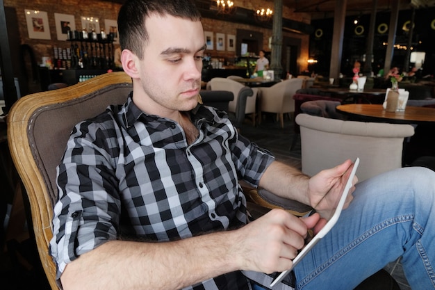 Een succesvolle jongeman in een donker geruit overhemd in het café doet zaken. jonge hipster die digitale tablet in wapens houdt. beambte op lunch.