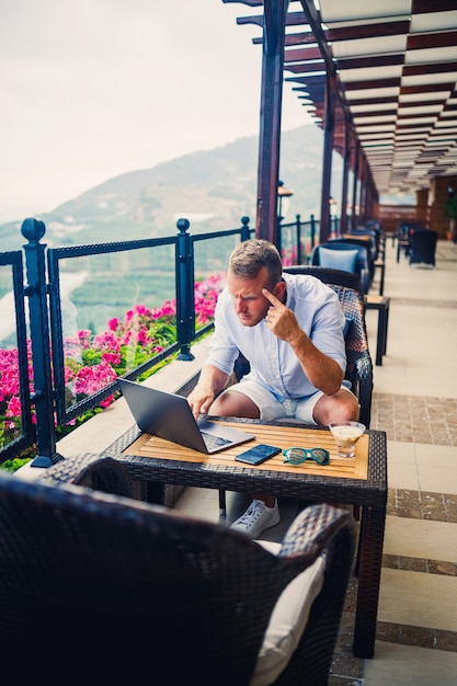 Een succesvolle mannelijke zakenman zit aan een tafel met een laptop hij werkt op vakantie Vakantie en werken op afstand De man zit in een restaurant op een terras met een panoramisch uitzicht