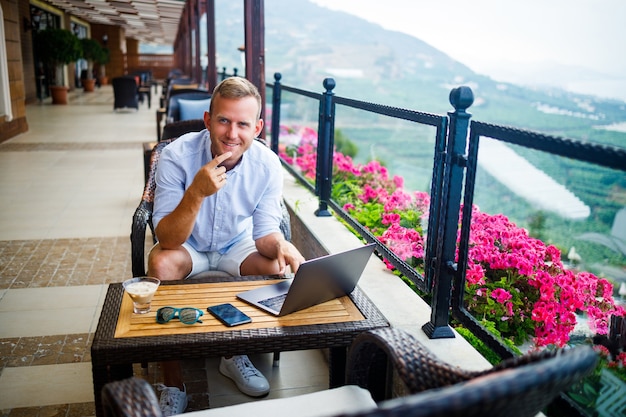 Een succesvolle mannelijke zakenman zit aan een tafel met een laptop, hij werkt op vakantie. Vakantie en werken op afstand. De man zit in een restaurant op een terras met panoramisch uitzicht