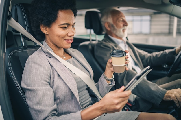 Een succesvolle zwarte zakenvrouw die digitale tablet gebruikt en koffie drinkt om te gaan terwijl ze met haar senior collega met de auto reist.