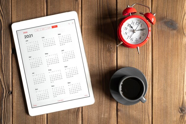 Een tablet met een open kalender voor het jaar 2021, een kopje koffie en een rode wekker op de achtergrond van een houten planken tafel