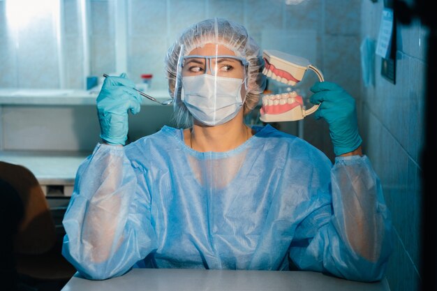 Een tandarts die blauwe handschoenen en een masker draagt, houdt een tandmodel van de boven- en onderkaak en een tandheelkundige spiegel vast