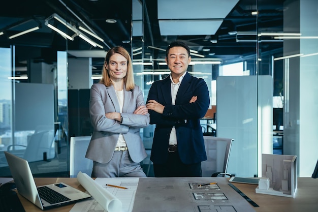 Een team van architecten van gemengd ras, een Aziatische man en een blanke vrouw die met succes glimlachen en naar de gekruiste cameraarmen kijken terwijl ze in een modern kantoor werken aan een tekening en huisplan