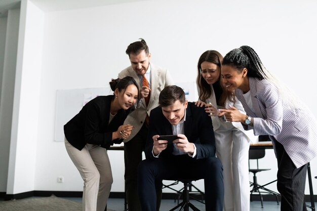 Een team van professionele kantoormedewerkers staat rond een collega die op een mobiele telefoon speelt in de