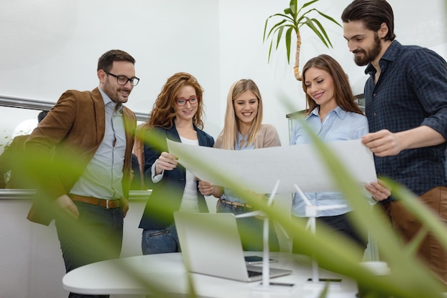 Een team van slimme gekwalificeerde bekwame jonge ingenieurs die discussiëren over het project van alternatieve energie met miniaturen van windmolenturbines aan tafel.