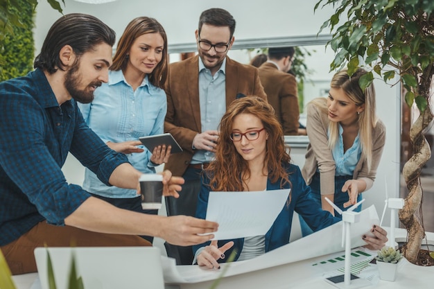 Een team van slimme gekwalificeerde, bekwame jonge ingenieurs discussieert over het project van alternatieve energie met miniaturen van windmolenturbines aan tafel.