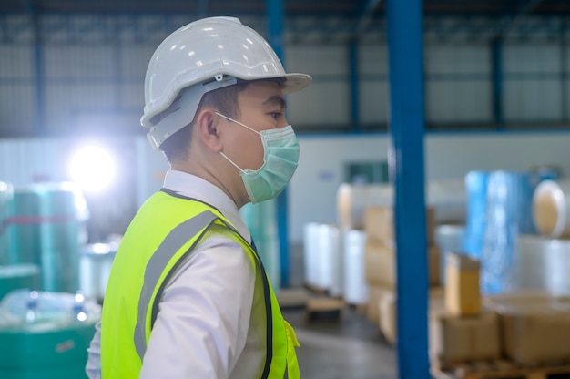 Een technische man met een medisch masker, een beschermende helm die in een magazijnfabriek werkt