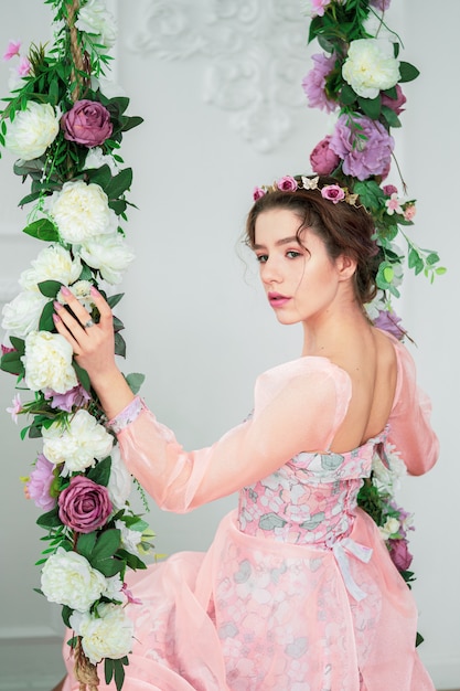 Een teder jong meisje naar het beeld van een prinses in een roze jurk met een krans op haar hoofd zit op een schommel met bloemen.
