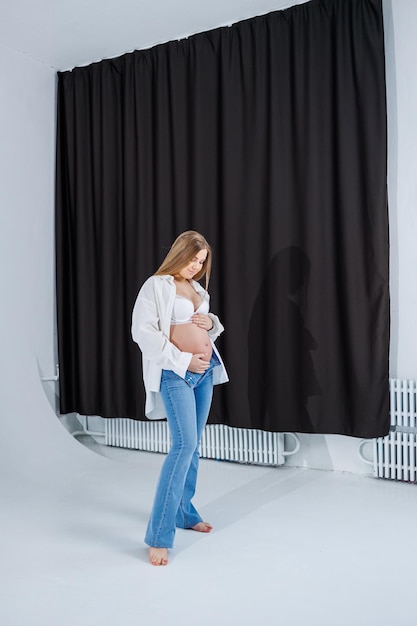 Een tedere jonge zwangere vrouw in een wit overhemd en spijkerbroek knuffels haar buik Zwangere gelukkige vrouw op een witte achtergrond