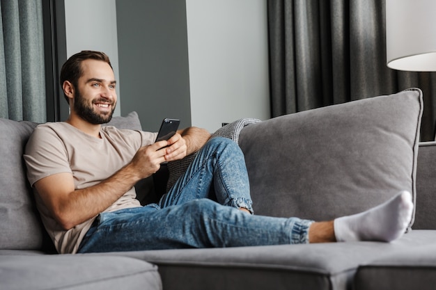 een tevreden gelukkige jonge man binnenshuis thuis op de bank met behulp van mobiele telefoon.