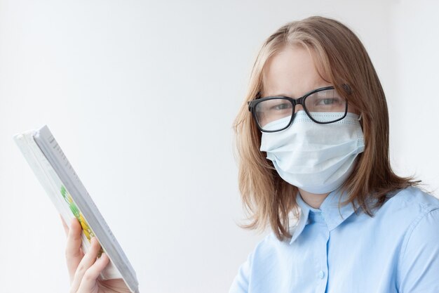 Een tienermeisje, blond, in een blauw shirt, een medisch masker en een bril op een witte achtergrond kijkt naar de camera en houdt een leerboek in haar hand.