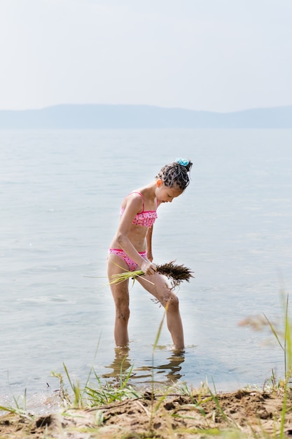 Een tienermeisje in een badpak waaiert haar dij uit met een bezem van gras terwijl ze in de zomer badprocedures nabootst op de zonnige oever van de rivier. Spelletjes en vrije tijd voor kinderen