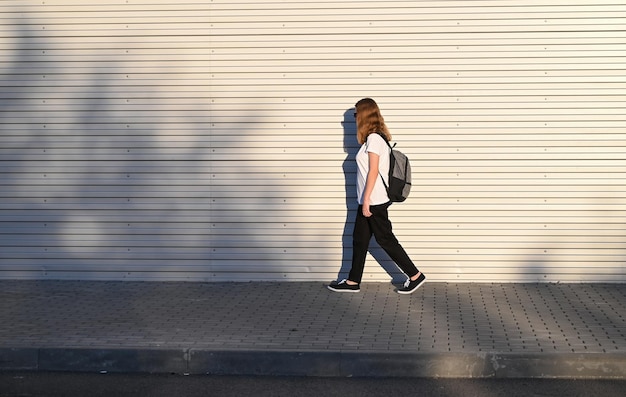 Een tienermeisje in een zwarte broek en een wit T-shirt loopt over straat met een zwarte rugzak op haar rug. Terug naar school