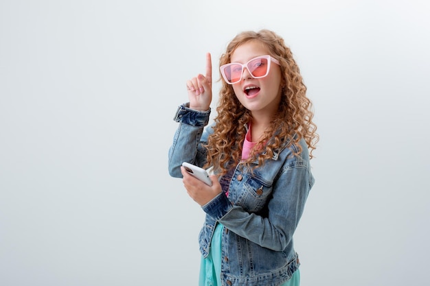 Een tienermeisje in zonnebril met een telefoon toont verrassing op een witte achtergrond