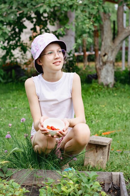 Een tienermeisje met een bril en een hoed met een kom aardbeien zit in de tuin