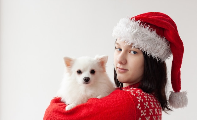 Een tienermeisje met een kerstmuts en een rode kersttrui houdt een witte pomeranian in haar armen. Het concept van Kerstmis en Nieuwjaar