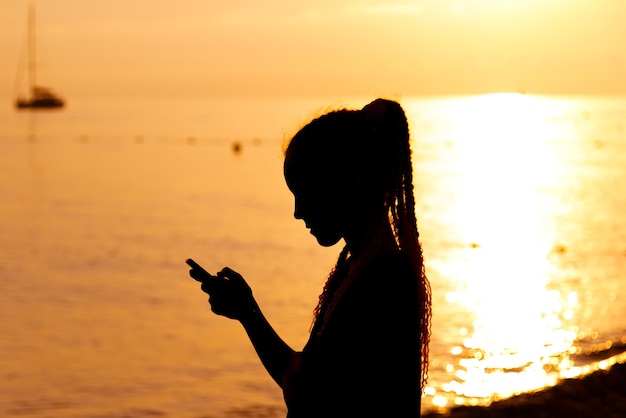 Een tienermeisje met een telefoon op de achtergrond van de zonsondergangzee Rust Communicatie online Zomer