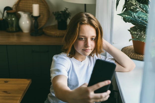 Een tienermeisje met rood haar zit bij het raam en kijkt in de telefoon