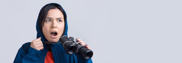 Een toeristenmeisje in een blauwe regenjas houdt een verrekijker in haar handen en kijkt in de verte spionnen