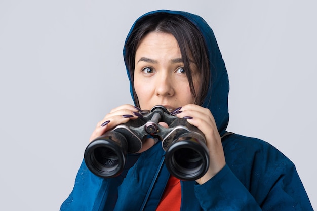 Een toeristenmeisje in een blauwe regenjas houdt een verrekijker in haar handen en kijkt in de verte, spionnen.