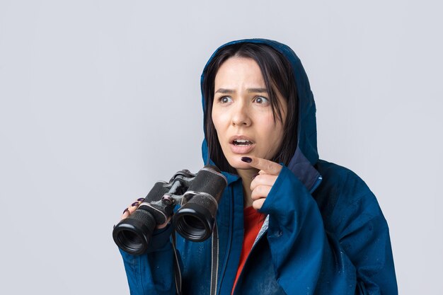 Een toeristenmeisje in een blauwe regenjas houdt een verrekijker in haar handen en kijkt in de verte spionnen