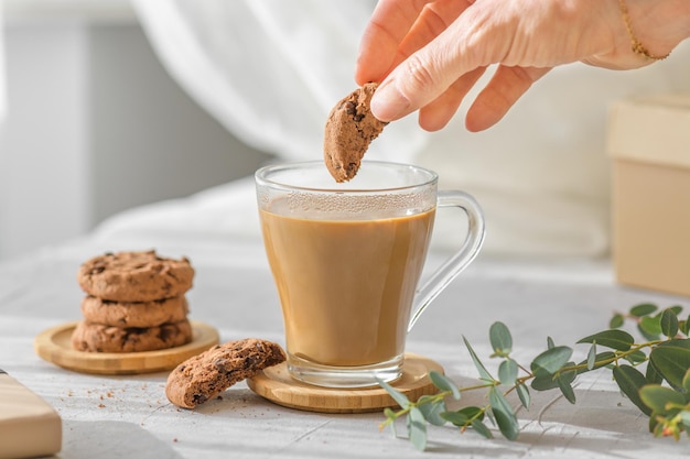 Een transparante kop koffie met melk en hand met chocolade koekjes eucalyptus bladeren in de ochtend schaduwen en zonnestralen door het raam op grijze tafel achtergrond Ochtendkoffie concept close-up