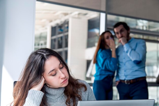 Een triest uitziende Turkse vrouw werkt aan een bureau en er zijn twee collega's die op de achtergrond achter haar aan het roddelen zijn