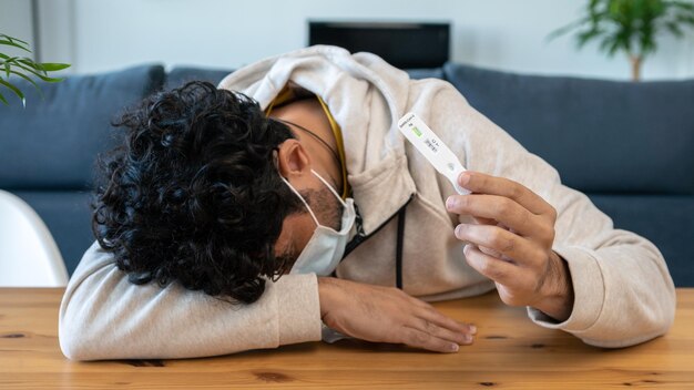 Een trieste blanke man maakt een snel thuistestapparaat voor het Covid19-virus met een positief resultaat en houdt zijn hoofd bij de hand aan een houten tafel Antigeencassette van pcr tegen nieuw Omicron-variantvirus