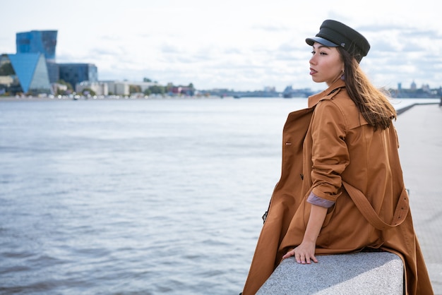 Een trieste eenzame vrouw met een Europees uiterlijk in een jas en een pet zit aan de oever van de rivier...