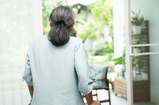 Een trieste oude oudere vrouw gebruikt walker om voor ramen te staan en naar buiten te kijken en zich eenzaam te voelen.