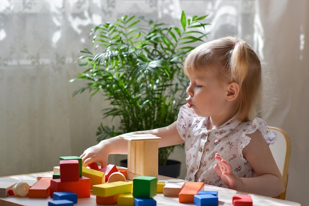 Een tweejarig meisje zit aan een tafel een houten bouwpakket in elkaar te zetten Een kind bouwt een toren van kubussen