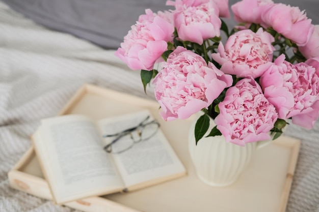 Een vaas met een boeket roze pioenrozen een opengeslagen boek en glazen op een dienblad op het bed