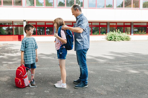 Foto een vader begeleidt zijn kinderen naar school en helpt zijn dochter een rugzak aan te trekken voor een schoolmeisje.