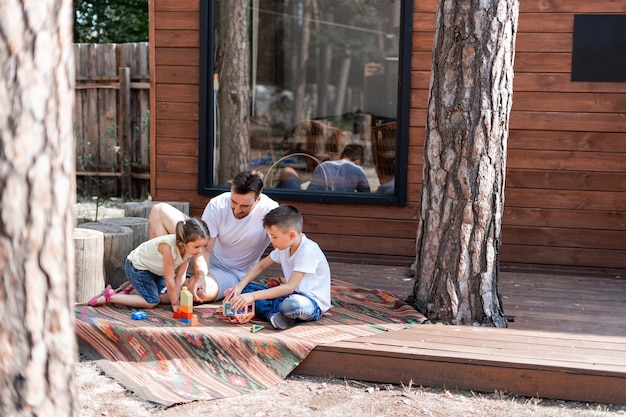 Een vader speelt met twee kinderen die op de veranda bij een houten landhuis zitten, besteedt zijn vrije tijd aan het helpen van kinderen om met speelgoed te spelen, brengt vakantie door met zijn gezin