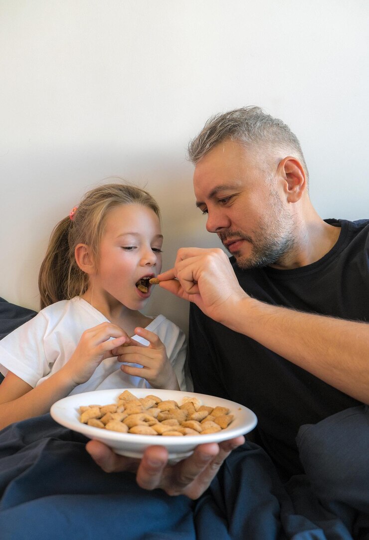 Een vader voedt zijn jonge dochter liefdevol met een gezonde snack in bed. Ze zijn geknuffeld onder een donkerblauw dekbed en creëren een warme en intieme sfeer. Dit beeld toont een liefdevolle familieverbinding.