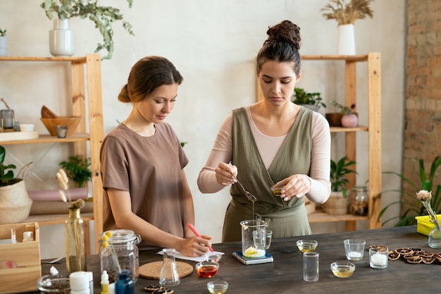 Een van de twee jonge vrouwen die etherische olie in glaswerk doen met gemalen zeepmassa terwijl ze een mengsel maken voor een natuurlijk, handgemaakt cosmetisch product