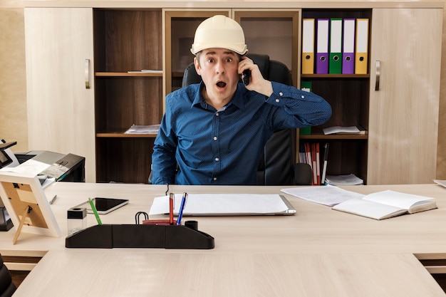 Een verbaasde baas met een bouwhelm aan de tafel in zijn kantoor roept iets in de telefoon. Belangrijk nieuws. Online communicatie. Zakenman.