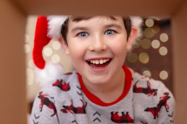 Een verbaasde, gelukkige brunette jongen in een nieuwjaarstrui en kerstman hoed neemt een cadeautje