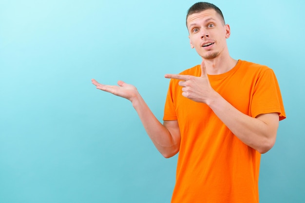 Een verbaasde man in een oranje t-shirt die met een hand wijst en met de vinger naar de bovenzijde wijst met lege kopie ruimte op blauwe achtergrond Knap vrolijk model aanwezig product