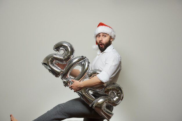 Een verbaasde man met een baard in een roodfluwelen kerstmuts houdt zilveren ballonnen vast in de vorm van 2022. Nieuwjaarsfeest.