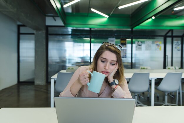 Een verdrietig meisje met een kopje koffie in haar handen gebruikt een laptop op het bureau in het kantoor.