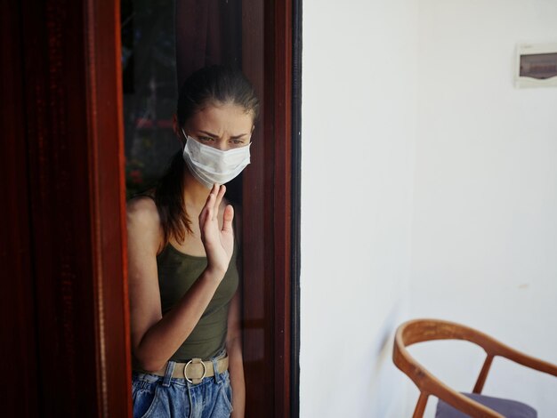 Een verdrietige vrouw met een medisch masker die uit het raam kijkt. Hoogwaardige foto.