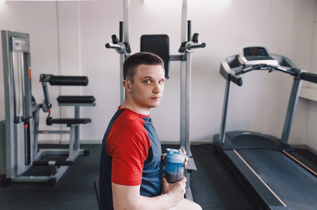 Een vermoeide atleet met een nat gezicht zit op een bankje in de sportschool met een shaker eiwit.